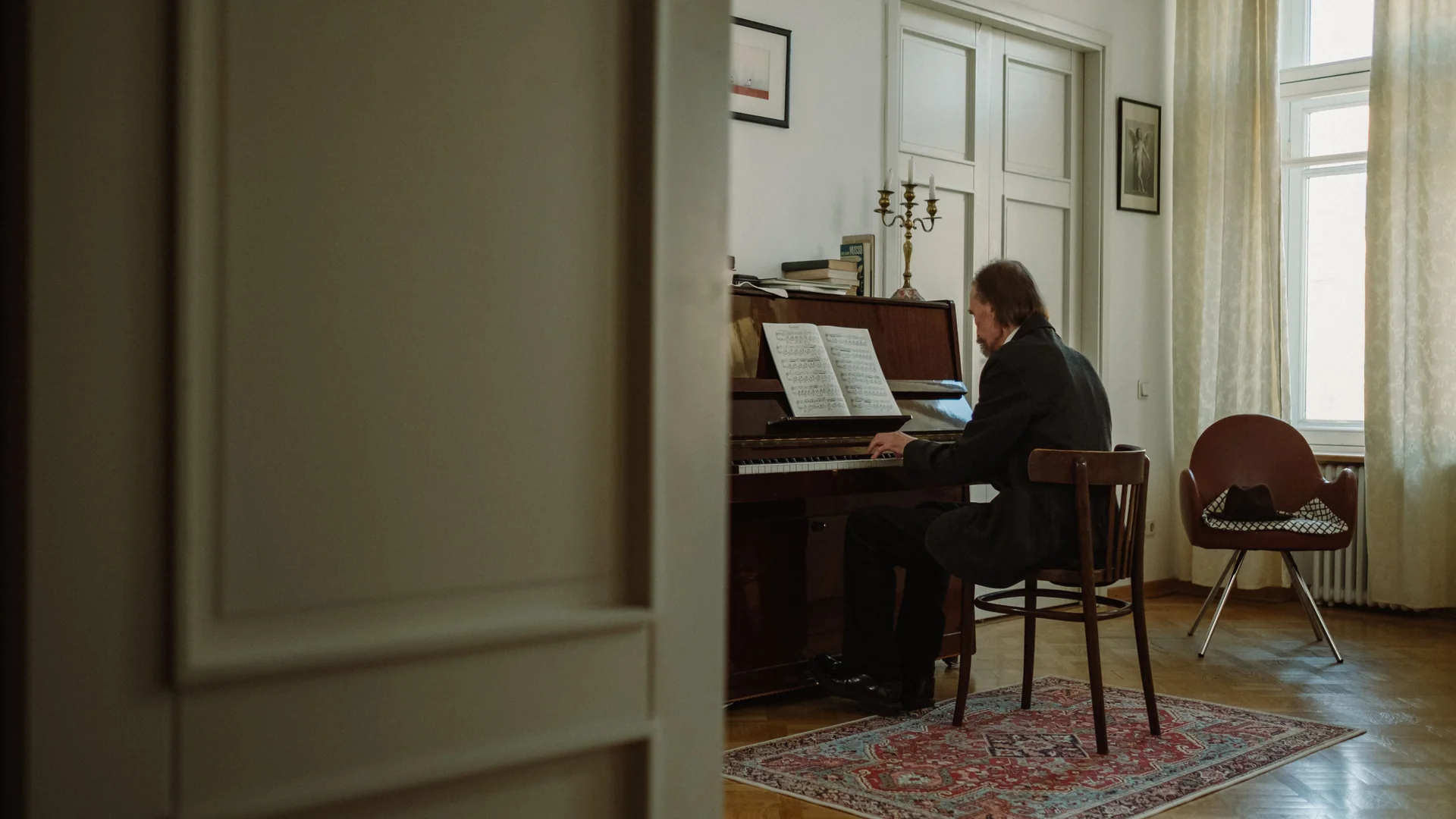 Ein Mann in dunklem Mantel spielt auf einem Klavier mit aufgeschlagener Partitur in einem hellen, hohen Raum mit Messing-Kandelaber, Parkettboden und hohen Fenstern (nicht der Chopin-Salon).