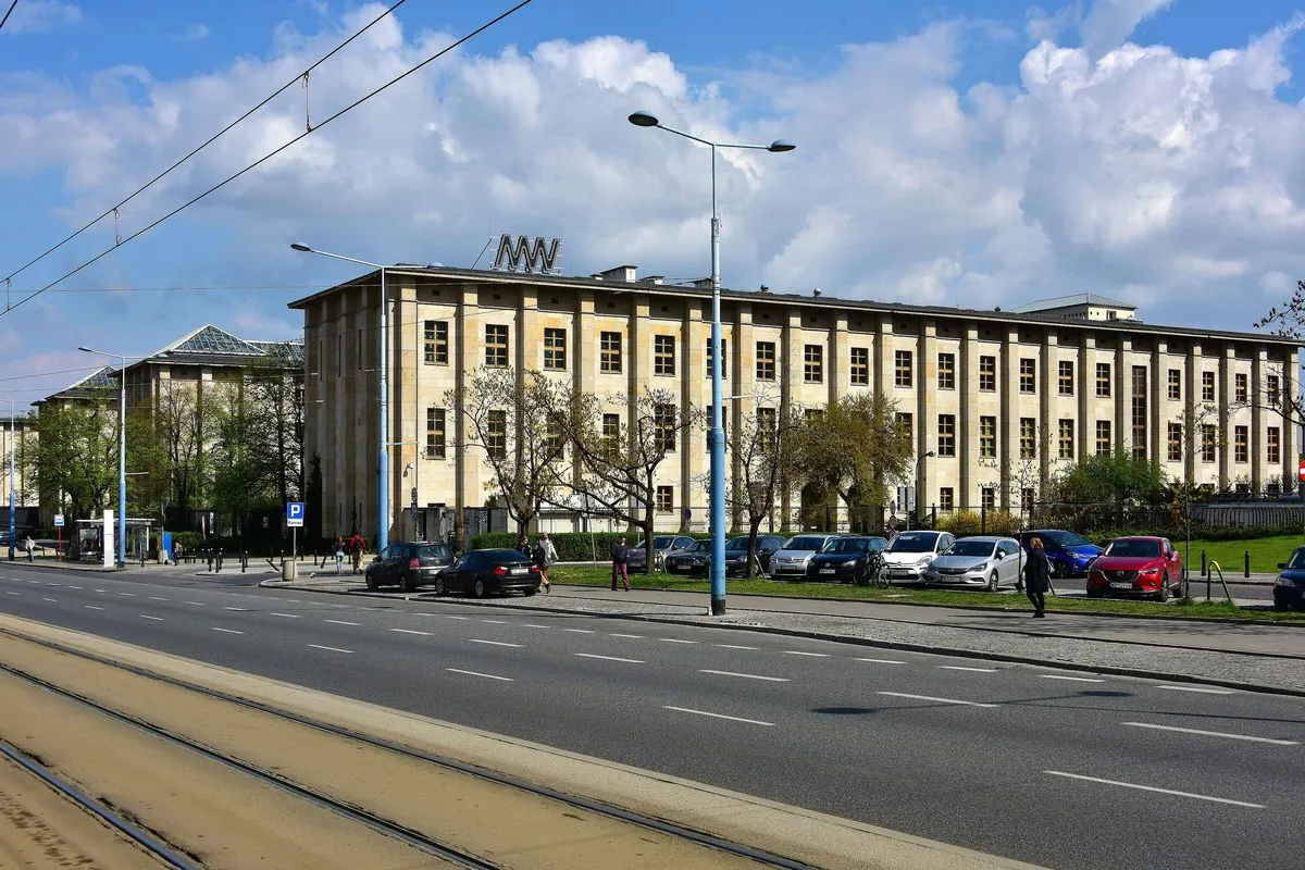 Das lange, helle steinerne Hauptgebäude des Nationalmuseums in Warschau, gesehen über eine breite Straße mit Straßenbahnschienen und Bäumen