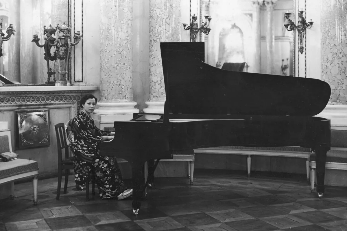 Eine Schwarz-Weiß-Fotografie einer Frau im Kimono, die in einem Säulensaal mit Kandelabern an den Wänden auf einem Flügel spielt
