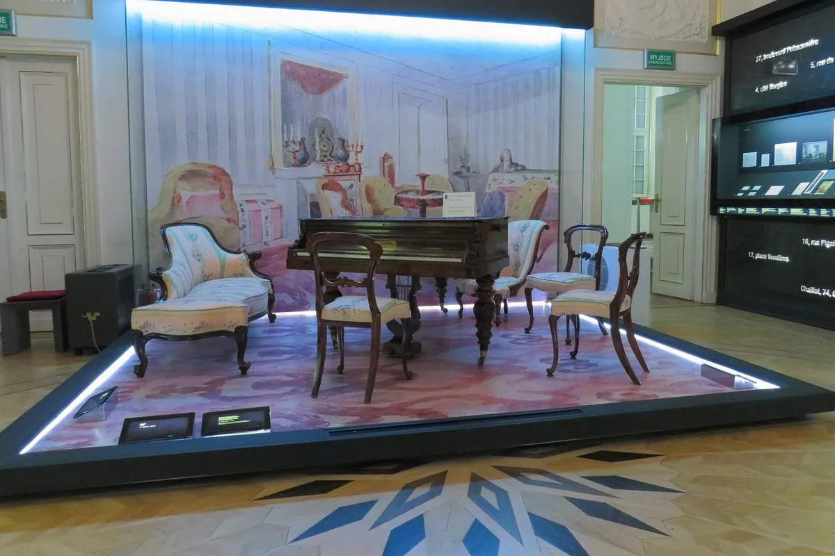 Ein kleiner Flügel mit einer Partitur auf dem Notenpult steht zwischen Stühlen und einem Sofa in einer beleuchteten Museumsvitrine, die wie ein Salon eingerichtet ist